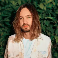 Kevin Parker, uno dei membri dei Tame Impala, con lunghi capelli castani e una camicia floreale, sullo sfondo di un muro di foglie verdi.
