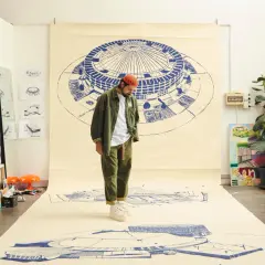 Un artista con un cappello arancione osserva disegni architettonici su carta, come se stesse creando nuove canzoni di Frah Quintale.