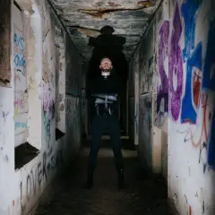 Uomo con barba in piedi in un corridoio pieno di graffiti, con indosso un abito nero. Sembra un rapper pronto per una sfida.