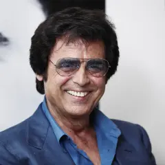 Un uomo sorridente con occhiali da sole aviator, indossa una giacca blu. Potrebbe essere Little Tony, pronto a cantare le sue canzoni.