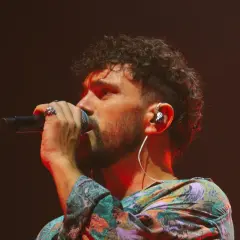 Marco Castello canta con passione, microfono in mano, cuffie nell'orecchio e un look colorato.