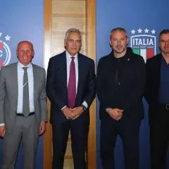 Quattro uomini in posa davanti a uno sfondo blu con il logo FIGC. Nessuno sembra un "dead can dance", ma tutti sono vestiti elegantemente.