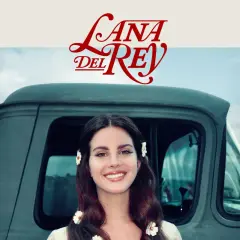 Lana Del Rey sorride, con fiori tra i capelli, accanto a un vecchio camion. Un'immagine che evoca il suo "lust for life".