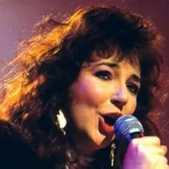 Kate Bush ora, con i suoi capelli scuri e fluenti, canta con passione al microfono, illuminata da luci calde.