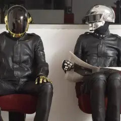 I Daft Punk, in pelle nera e caschi iconici, uno dorato e l'altro argentato, siedono leggendo il giornale.