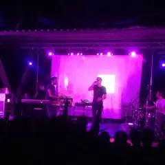 Concerto con luci viola. Sul palco, un cantante con occhiali e una tastiera. Dietro, un batterista e altri musicisti. Il pubblico è in ombra.