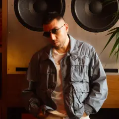 Man in sunglasses and a grey jacket leans against a large speaker.