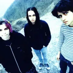 Three young people with dark hair and casual clothing stand outdoors in a rocky, wooded area.