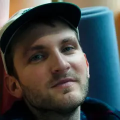 Ritratto di un uomo con cappellino, circondato da rotoli di gommapiuma colorata. Le sue canzoni di Giorgio Poi evocano immagini vivide.