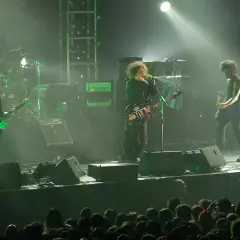 Sul palco, i membri dei The Cure suonano le loro iconiche canzoni. Un chitarrista con una chitarra verde brillante, il cantante con la sua chitarra nera e un altro chitarrista.