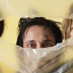 Occhi curiosi di Alberto Ferrari Verdena e altri due volti emergono da un telo di plastica trasparente su sfondo giallo.