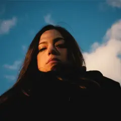 Daniela Pes spira sotto un cielo azzurro e nuvoloso, con uno sguardo intenso e capelli scuri che incorniciano il viso.