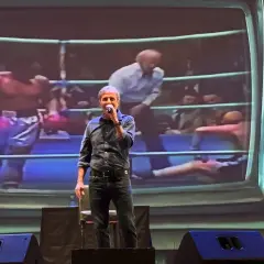 Uomo sul palco con microfono, sullo sfondo un ring di pugilato proiettato. "Dai mamma dai", urla la folla.