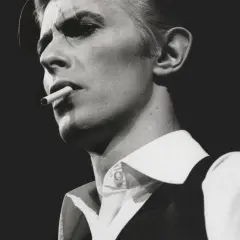David Bowie, un'icona dei suoi golden years, produttore di musica e stile, fuma una sigaretta con sguardo intenso.