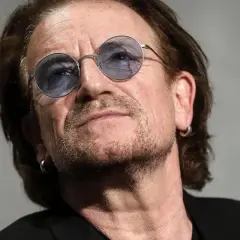 Primo piano di Bono Vox con occhiali rotondi blu, che ricorda le sue iconiche canzoni.