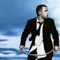 Copertina album di Eros Ramazzotti con lui in giacca e cravatta su una bicicletta sotto un cielo blu.
