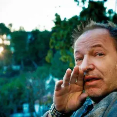 Vasco Rossi sussurra "ti prendo e ti porto via" con la mano all'orecchio, mentre il sole tramonta tra gli alberi.