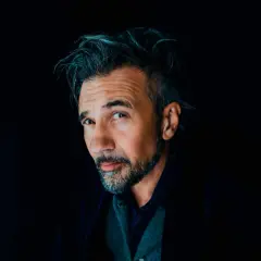 Ritratto intenso di John de Leo, con capelli spettinati e sguardo penetrante, su sfondo scuro.