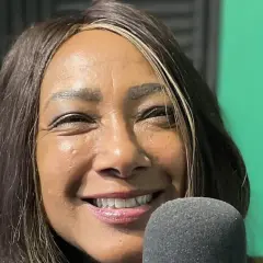 Dee C. Lee sorride vicino a un microfono, con uno sfondo verde e pannelli acustici.