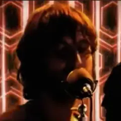 Un uomo con capelli castani canta al microfono, sullo sfondo un motivo esagonale rosso. Sembra che baustelle charlie fa surf con la sua musica.