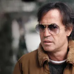 Close-up of a man with dark hair, wearing aviator sunglasses and a brown jacket with a fur collar.
