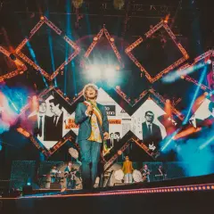 Cantante sul palco con sfondo di immagini e luci. I testi di Vasco Rossi sono ancora in coma, ma la musica continua.
