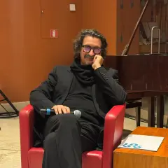 Sergio Cammariere, con microfono in mano, siede su una poltrona rossa, pronto a raccontare le sue canzoni.