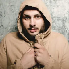 Uomo con cappuccio beige, sguardo intenso, barba. Sembra che qualcuno abbia sbagliato con lui.