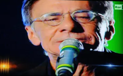 Roberto Vecchioni canta "Chiamami ancora amore" con passione, il microfono vicino alle labbra, sullo sfondo verde di Rai 1.