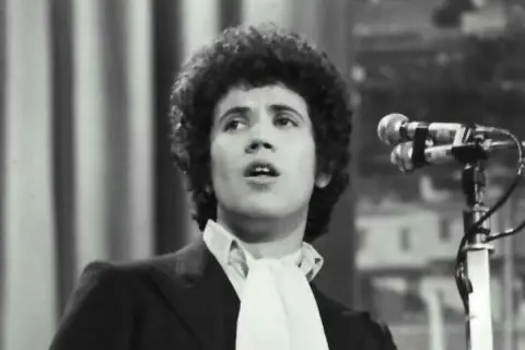 Lucio Battisti, con la sua iconica chioma, canta "Il tempo di morire" davanti a un microfono.