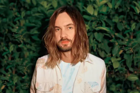 Kevin Parker, uno dei membri dei Tame Impala, con lunghi capelli castani e una camicia floreale, sullo sfondo di un muro di foglie verdi.