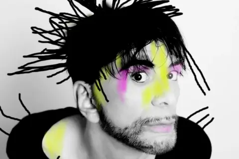Ritratto di Ivan Cattaneo con capelli neri a punta, trucco colorato e barba. Le sue canzoni sono uniche.
