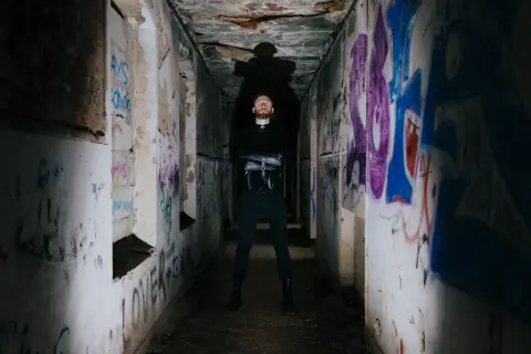 Uomo con barba in piedi in un corridoio pieno di graffiti, con indosso un abito nero. Sembra un rapper pronto per una sfida.