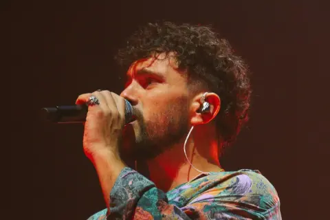 Marco Castello canta con passione, microfono in mano, cuffie nell'orecchio e un look colorato.