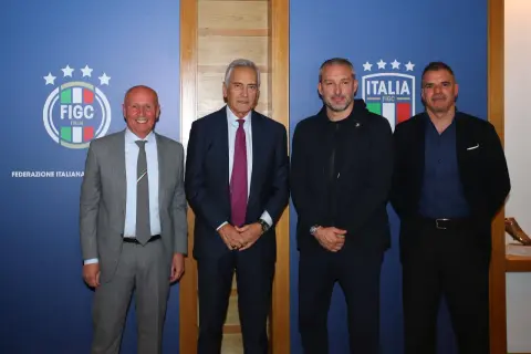 Quattro uomini in posa davanti a uno sfondo blu con il logo FIGC. Nessuno sembra un "dead can dance", ma tutti sono vestiti elegantemente.