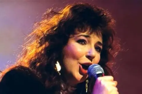 Kate Bush ora, con i suoi capelli scuri e fluenti, canta con passione al microfono, illuminata da luci calde.