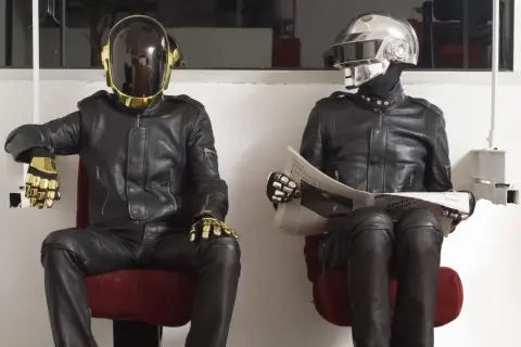 I Daft Punk, in pelle nera e caschi iconici, uno dorato e l'altro argentato, siedono leggendo il giornale.