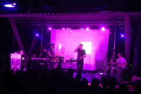 Concerto con luci viola. Sul palco, un cantante con occhiali e una tastiera. Dietro, un batterista e altri musicisti. Il pubblico è in ombra.