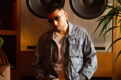 Man in sunglasses and a grey jacket leans against a large speaker.
