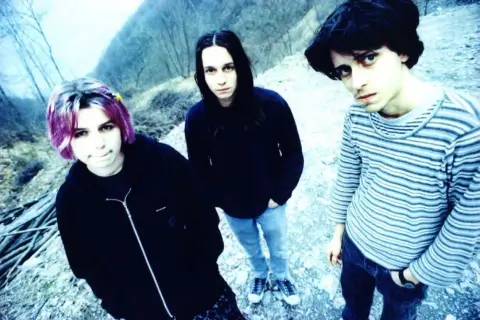 Three young people with dark hair and casual clothing stand outdoors in a rocky, wooded area.
