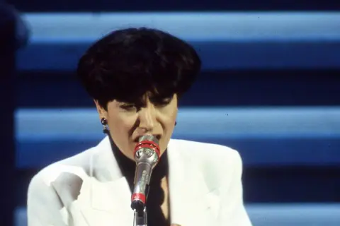 Mia Martini, icona della musica italiana, canta con passione sul palco, illuminata da luci soffuse.