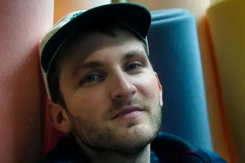 Ritratto di un uomo con cappellino, circondato da rotoli di gommapiuma colorata. Le sue canzoni di Giorgio Poi evocano immagini vivide.