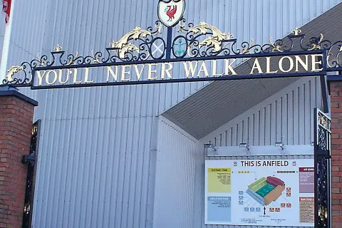 L'iconico cancello di Anfield con la scritta "You'll Never Walk Alone" e lo stemma del Liverpool. Un simbolo di unità e passione.