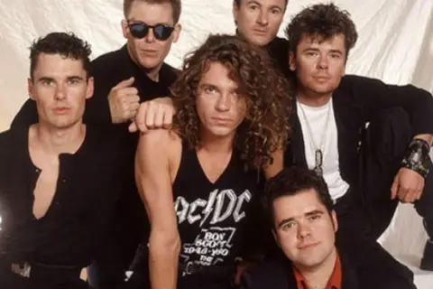 I membri degli INXS posano insieme, un gruppo di sei uomini con stili diversi, pronti a conquistare il mondo con la loro musica.