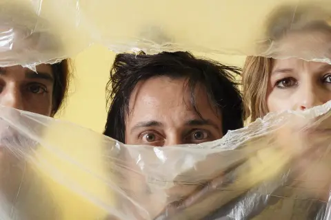 Occhi curiosi di Alberto Ferrari Verdena e altri due volti emergono da un telo di plastica trasparente su sfondo giallo.