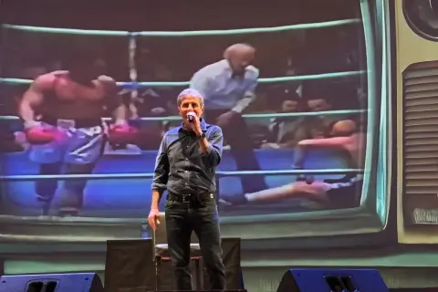 Uomo sul palco con microfono, sullo sfondo un ring di pugilato proiettato. "Dai mamma dai", urla la folla.