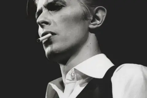 David Bowie, un'icona dei suoi golden years, produttore di musica e stile, fuma una sigaretta con sguardo intenso.