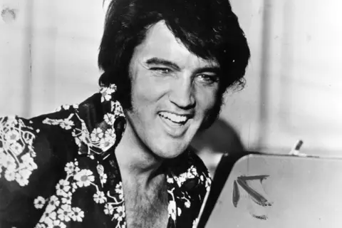Un giovane Elvis Presley, con la sua iconica capigliatura e una camicia floreale, sorride. Un'immagine che cattura un momento della sua storia.