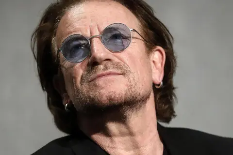 Primo piano di Bono Vox con occhiali rotondi blu, che ricorda le sue iconiche canzoni.