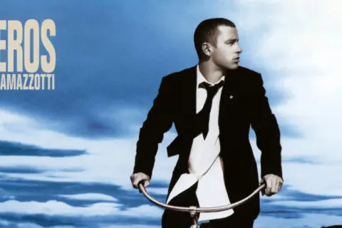 Copertina album di Eros Ramazzotti con lui in giacca e cravatta su una bicicletta sotto un cielo blu.